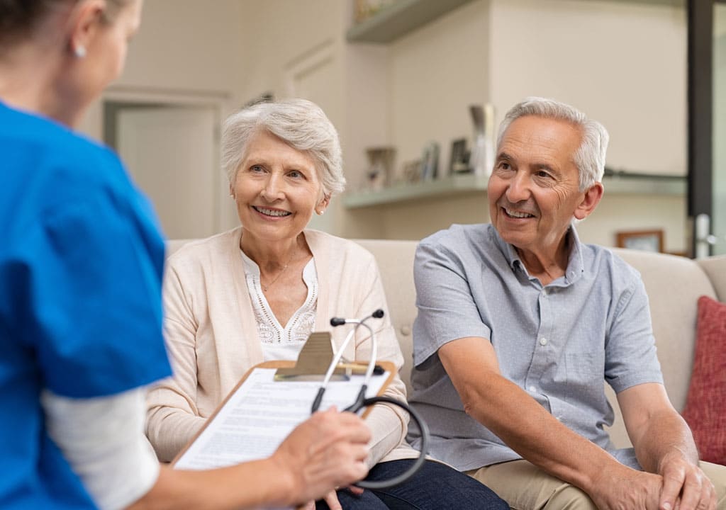Nurse during home care visit with senior couple. Nurse holding clipboard and stethoscope in conversation with old couple at home.